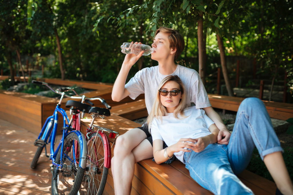 Young couple spending time on wood bench in park with two bicycles nearby. Boy sitting on bench and drinking water with beautiful girl leaning on him while lying on bench